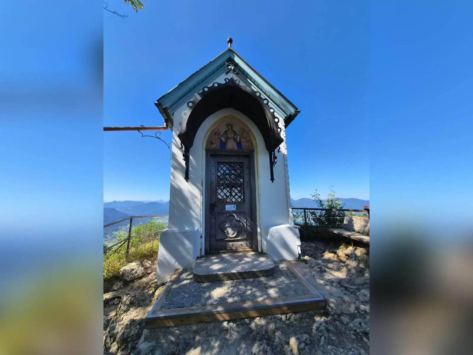 Kleine Kapelle auf dem Riederstein mit verziertem Eingang vor blauem Himmel; seitlich reicht der Blick in die Berglandschaft. | © DAV Markt Schwaben/Hubert Inhofer