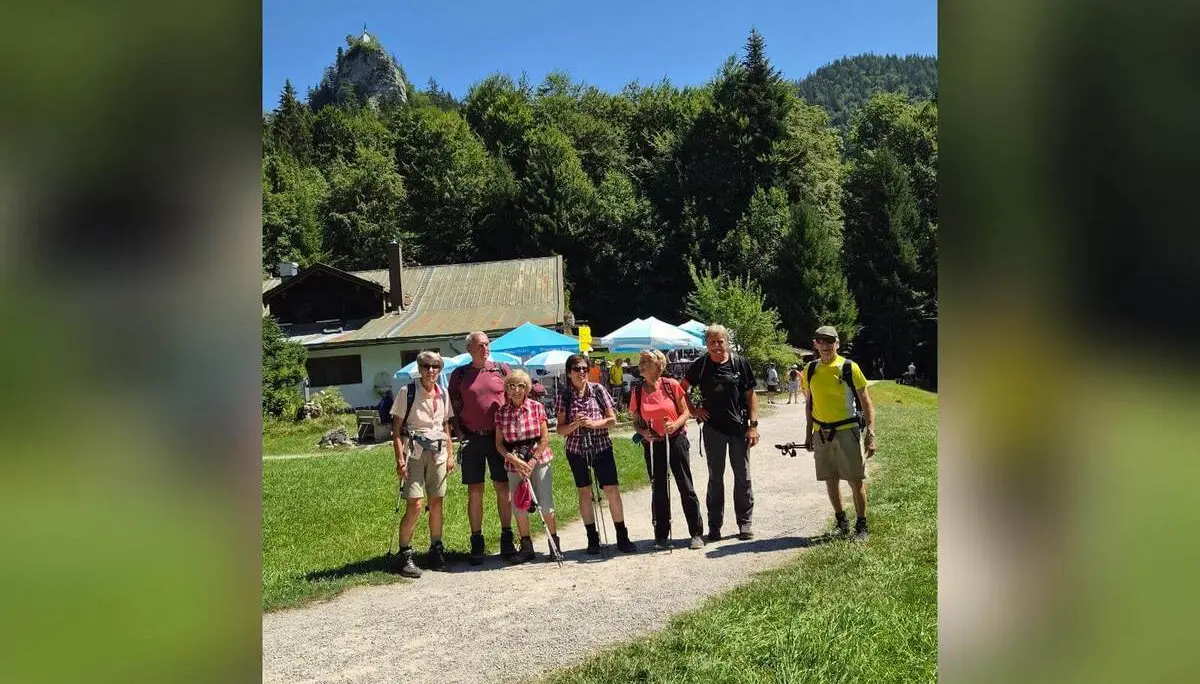 Wandergruppe steht vor einem Berggasthof auf einer Wiese; darüber erhebt sich der felsige Gipfel des Riederstein mit Kapelle. | © DAV Markt Schwaben/Hubert Inhofer