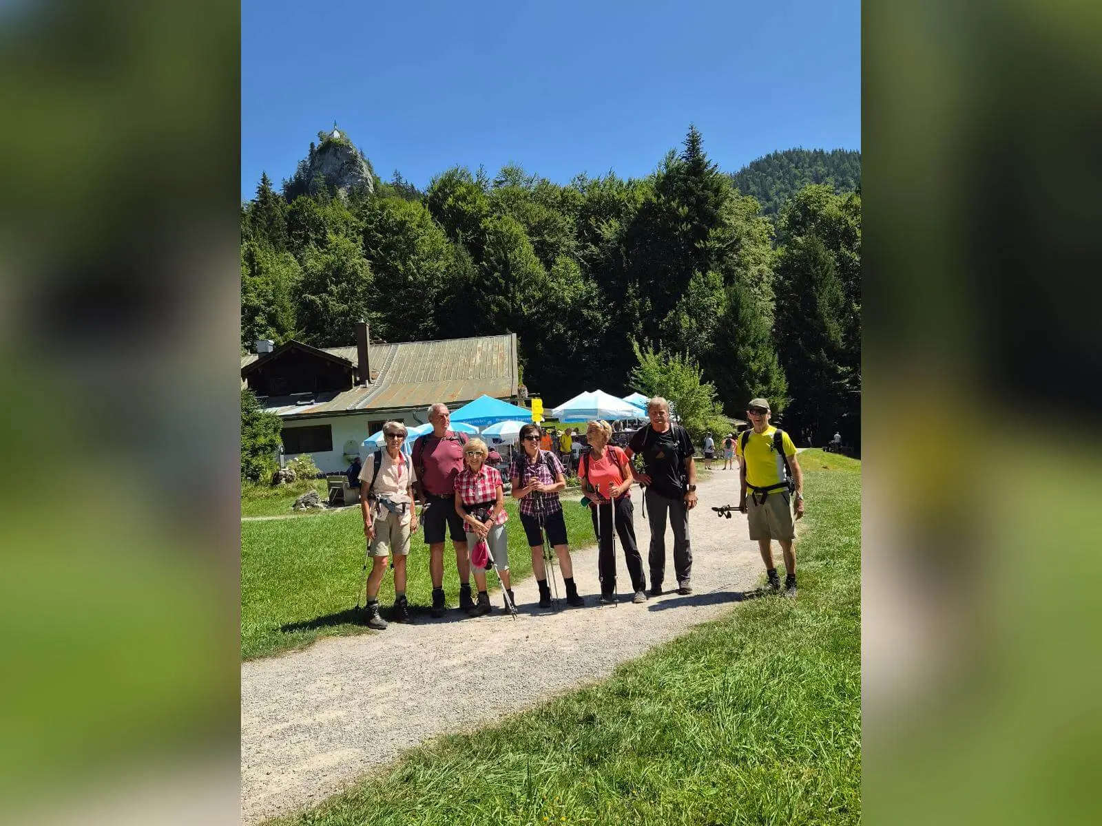 Wandergruppe steht vor einem Berggasthof auf einer Wiese; darüber erhebt sich der felsige Gipfel des Riederstein mit Kapelle. | © DAV Markt Schwaben/Hubert Inhofer