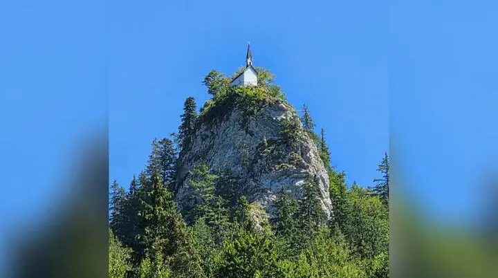 Die Riedersteinkapelle steht auf einem markanten Felsgipfel über dem Wald und hebt sich vor wolkenlosem blauem Himmel ab. | © DAV Markt Schwaben/Hubert Inhofer