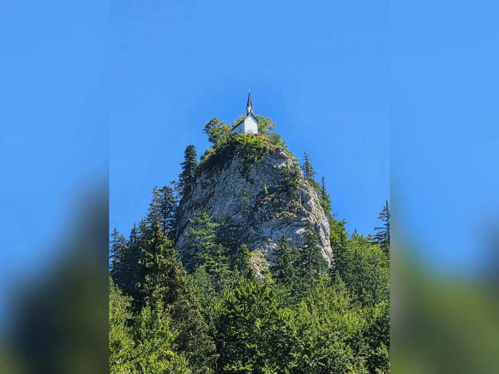 Die Riedersteinkapelle steht auf einem markanten Felsgipfel über dem Wald und hebt sich vor wolkenlosem blauem Himmel ab. | © DAV Markt Schwaben/Hubert Inhofer