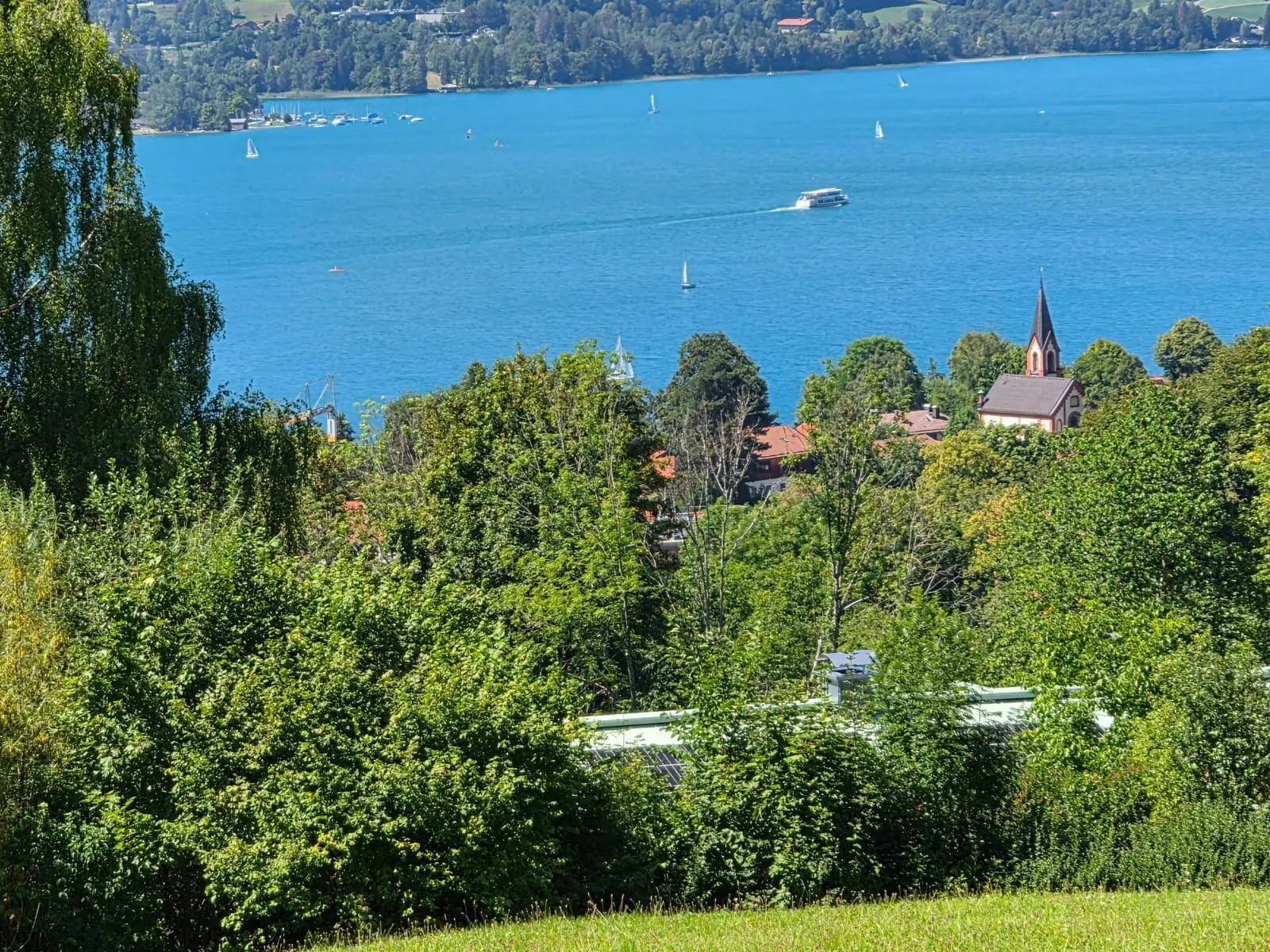 Blick über Baumwipfel auf den Tegernsee mit Segelbooten und Ausflugsschiff; zwischen den Bäumen ragt ein Kirchturm hervor. | © DAV Markt Schwaben/Hubert Inhofer
