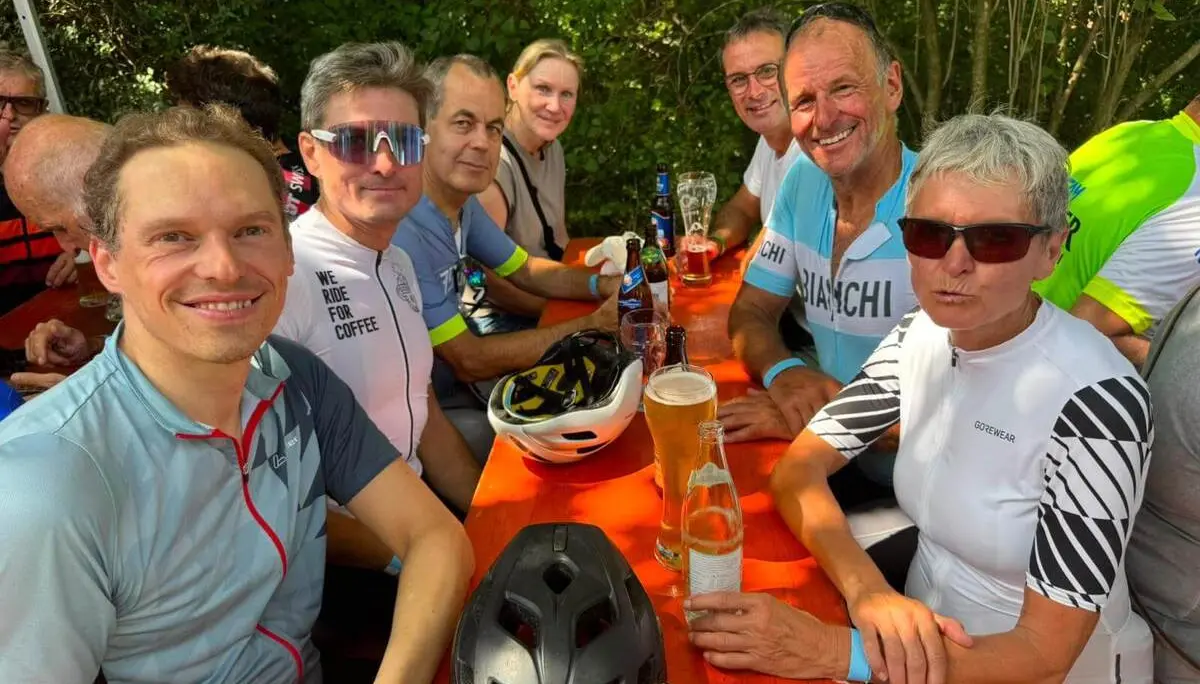 Mehrere Teilnehmende der Radtour sitzen an einem langen Holztisch im Freien. Auf dem Tisch stehen Getränke und Fahrradhelme. | © DAV Markt Schwaben/Radsportgruppe