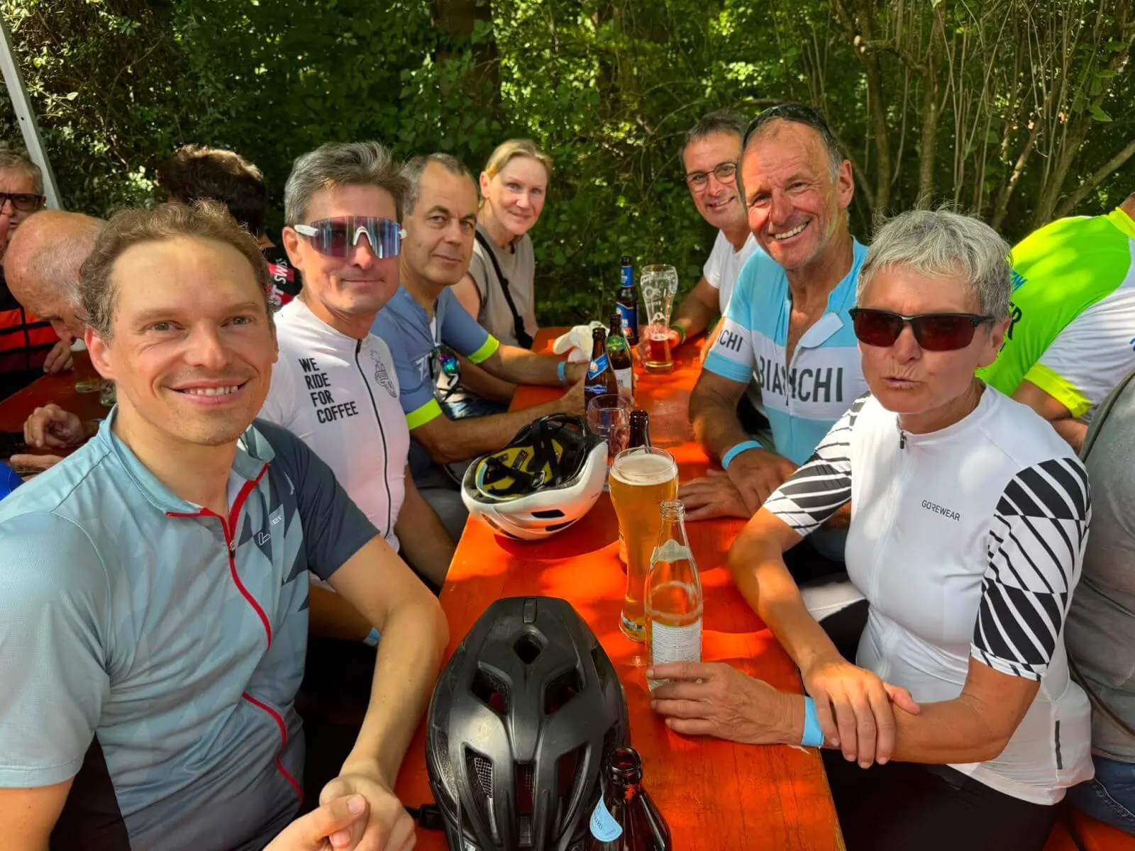 Mehrere Teilnehmende der Radtour sitzen an einem langen Holztisch im Freien. Auf dem Tisch stehen Getränke und Fahrradhelme. | © DAV Markt Schwaben/Radsportgruppe