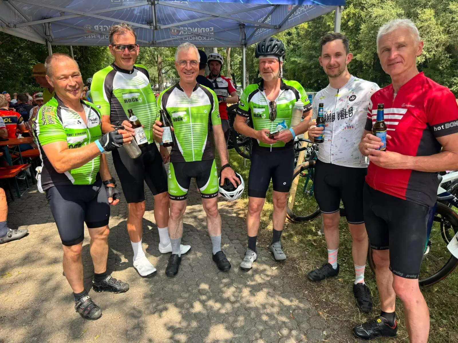 Sechs Teilnehmende der RTF stehen mit Getränken unter einem Erdinger-Alkoholfrei-Zelt. Im Hintergrund sind weitere Radfahrende und Rennräder zu sehen. | © DAV Markt Schwaben/Radsportgruppe