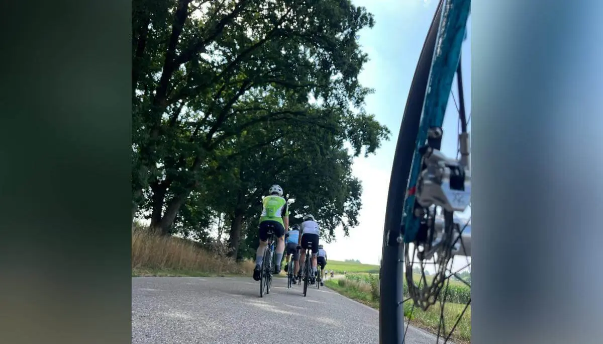 Mehrere Rennradfahrende fahren auf einer Landstraße durch die Landschaft. Im Vordergrund ist das Vorderrad eines Fahrrads aus niedriger Perspektive zu sehen. | © DAV Markt Schwaben/Radsportgruppe