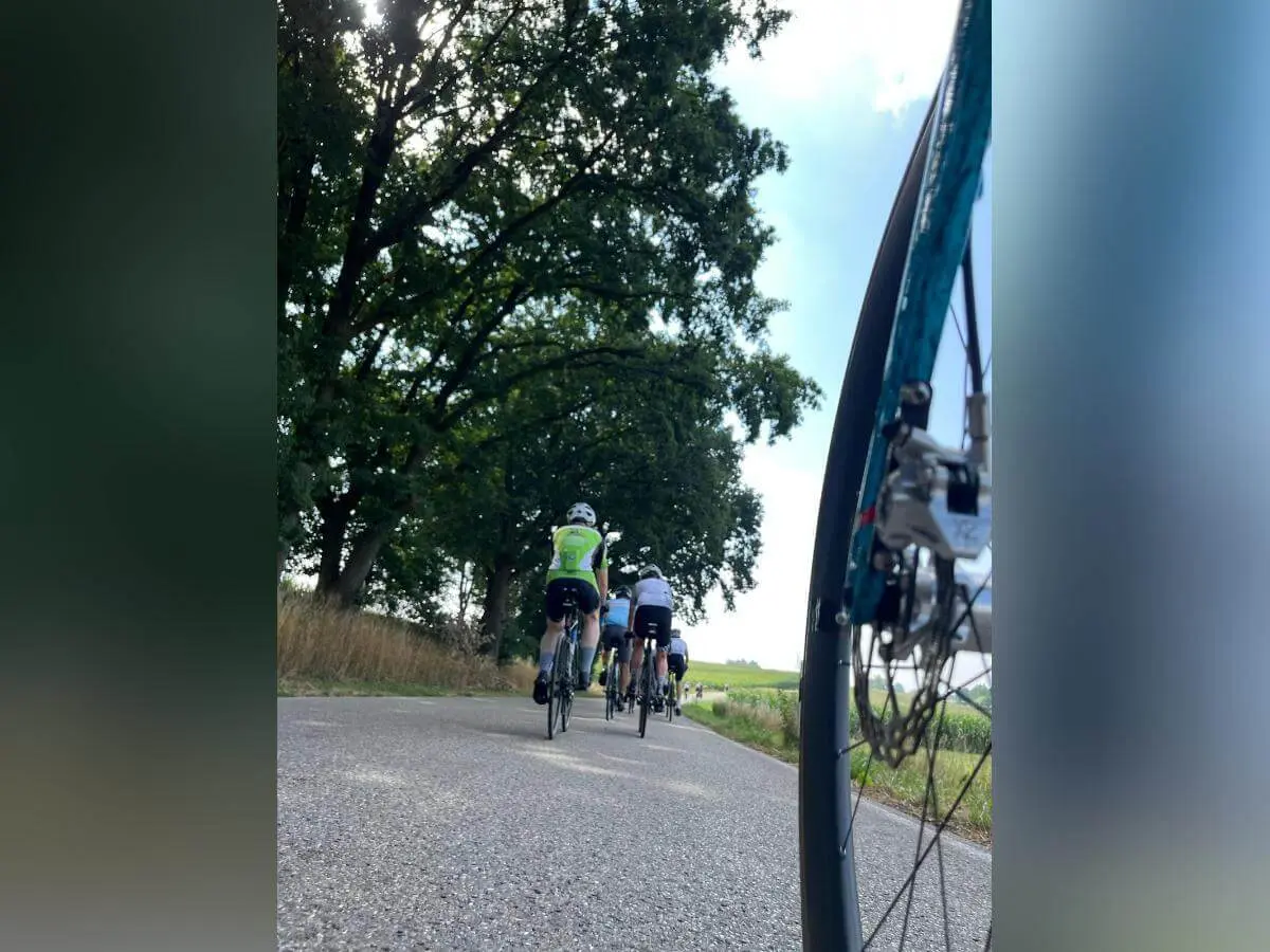 Mehrere Rennradfahrende fahren auf einer Landstraße durch die Landschaft. Im Vordergrund ist das Vorderrad eines Fahrrads aus niedriger Perspektive zu sehen. | © DAV Markt Schwaben/Radsportgruppe