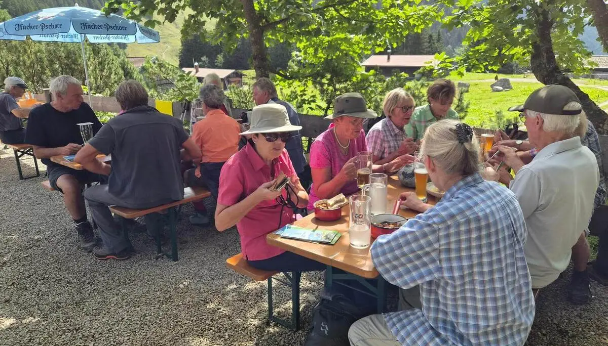 Mehrere Wandernde sitzen an Biertischen im Freien und essen. Im Hintergrund sind Almwiesen, Bäume und Hüttengebäude zu sehen. | © DAV Markt Schwaben/Hubert Inhofer