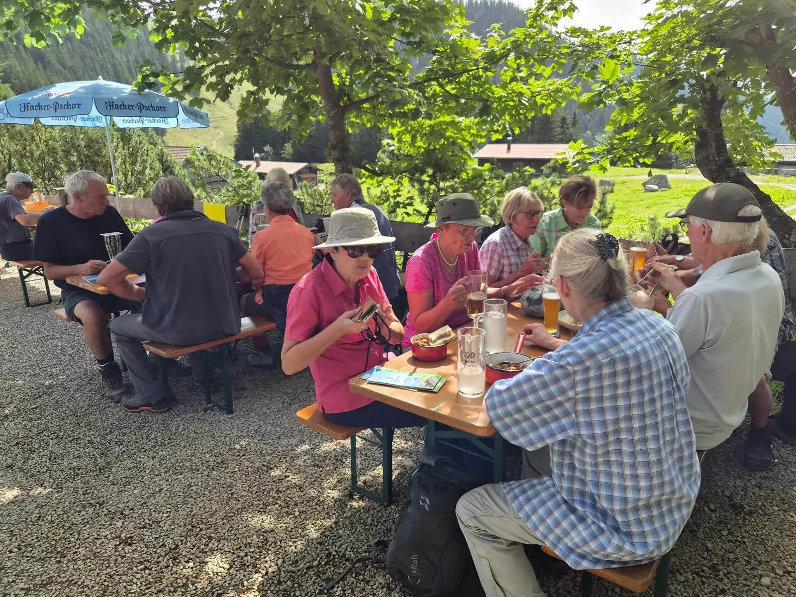 Mehrere Wandernde sitzen an Biertischen im Freien und essen. Im Hintergrund sind Almwiesen, Bäume und Hüttengebäude zu sehen. | © DAV Markt Schwaben/Hubert Inhofer
