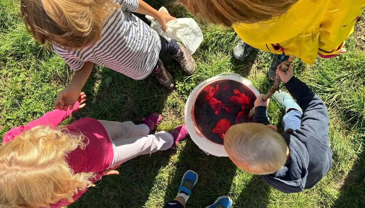 Mehrere Kinder stehen auf einer Wiese um einen Eimer mit rotem Farbbad und färben gemeinsam vorbereitete Kleidung zum Batiken ein. | © DAV Markt Schwaben / Lara Wolff