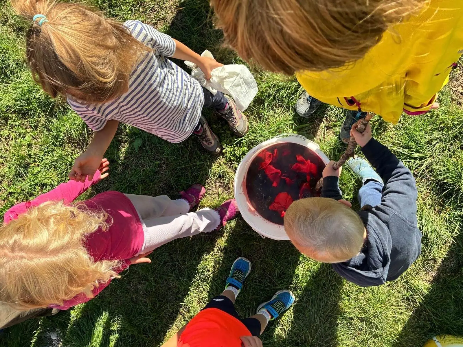 Mehrere Kinder stehen auf einer Wiese um einen Eimer mit rotem Farbbad und färben gemeinsam vorbereitete Kleidung zum Batiken ein. | © DAV Markt Schwaben / Lara Wolff