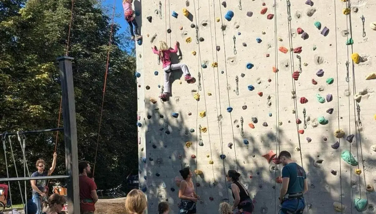 Familiengruppe an einer Outdoor-Kletterwand. Kinder klettern an der Wand, während weitere Teilnehmende unten sichern und zuschauen. | © DAV Markt Schwaben / Lara Wolff