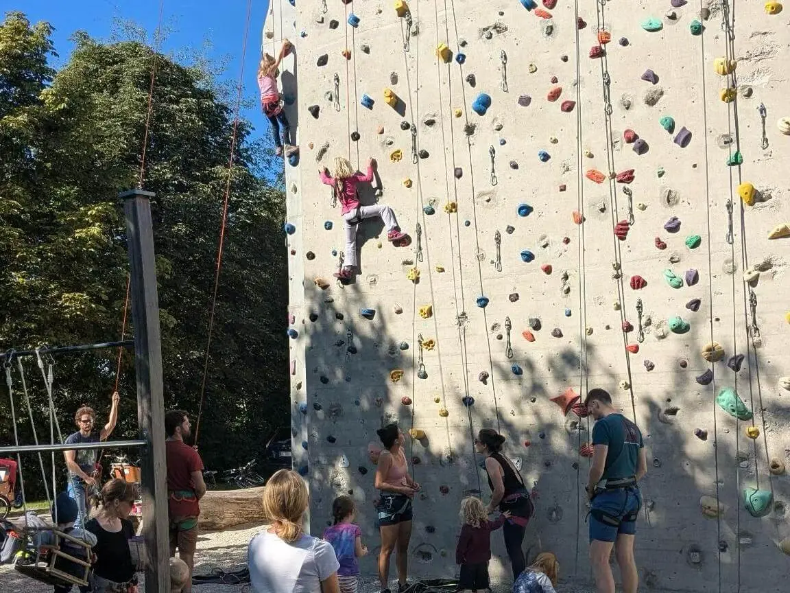 Familiengruppe an einer Outdoor-Kletterwand. Kinder klettern an der Wand, während weitere Teilnehmende unten sichern und zuschauen. | © DAV Markt Schwaben / Lara Wolff