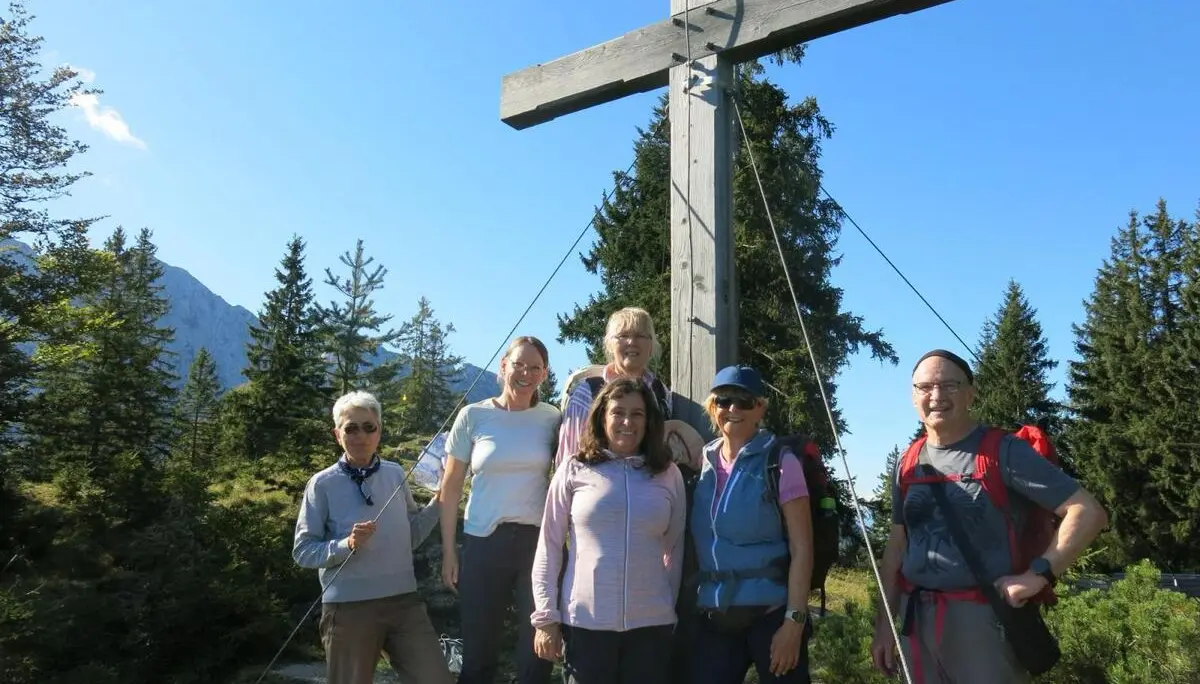 Sechs Wandernde stehen vor einem großen Gipfelkreuz auf dem Gamskogel. Dahinter erheben sich Nadelbäume und die Berge des Wilden Kaisers bei klarem Wetter. | © DAV Markt Schwaben / Erwin Matzinger