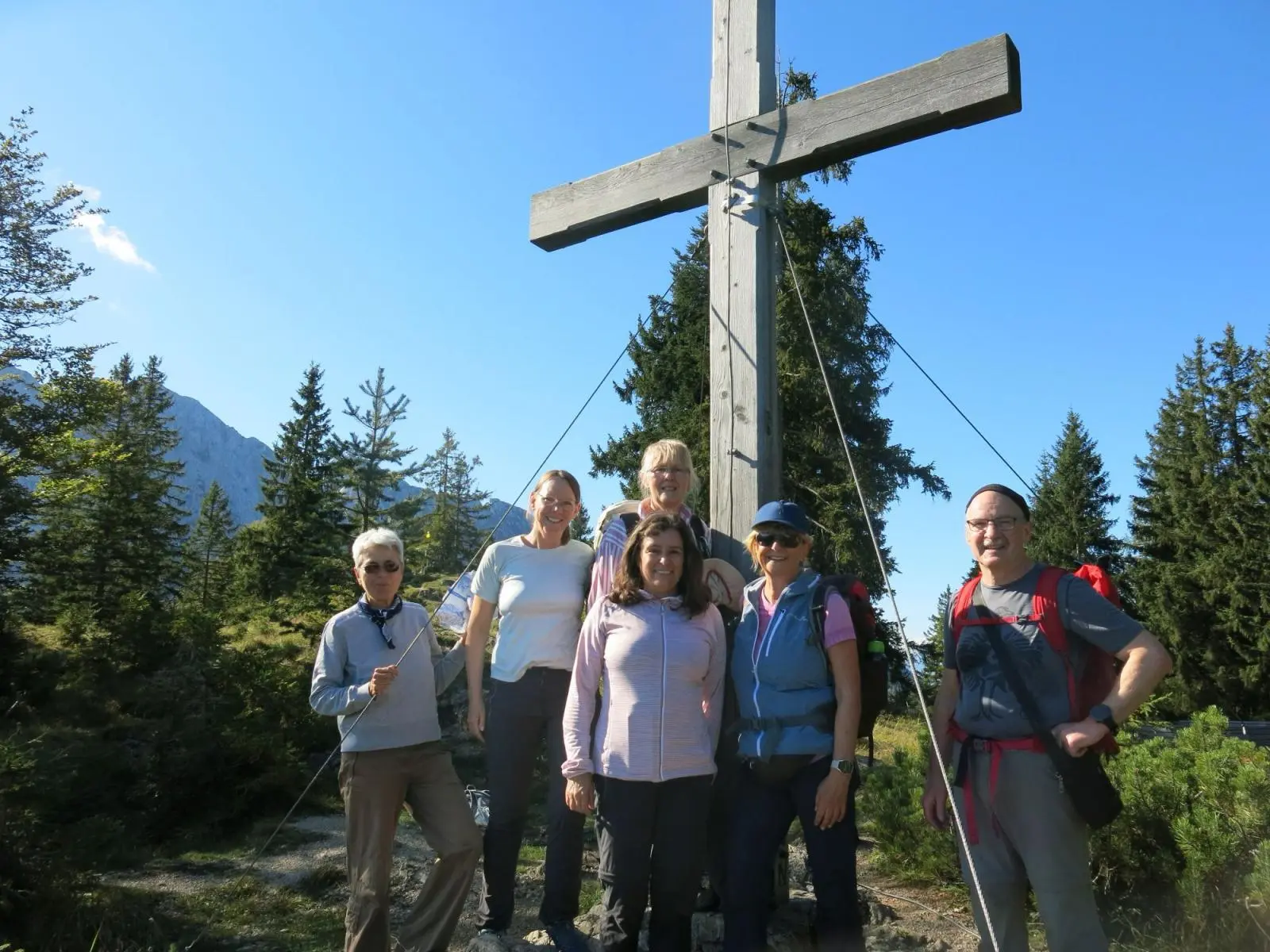 Sechs Wandernde stehen vor einem großen Gipfelkreuz auf dem Gamskogel. Dahinter erheben sich Nadelbäume und die Berge des Wilden Kaisers bei klarem Wetter. | © DAV Markt Schwaben / Erwin Matzinger