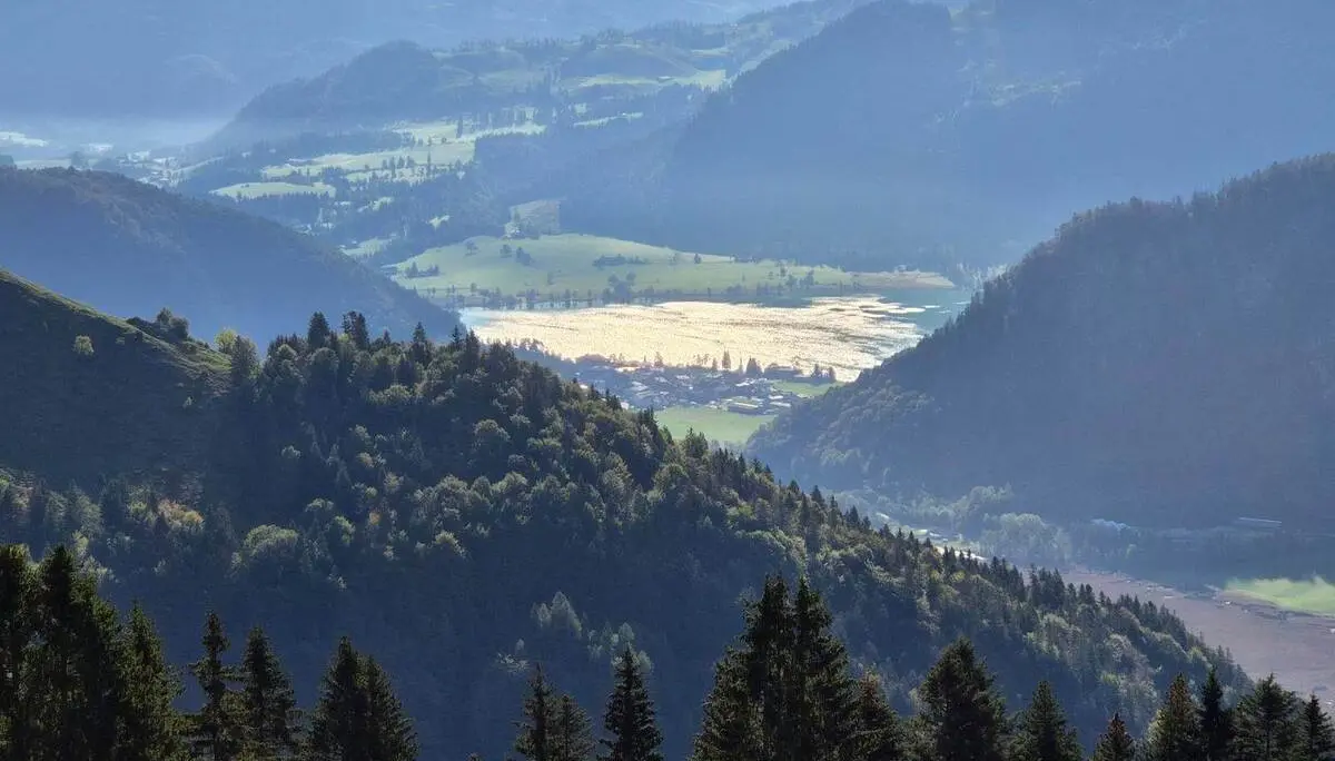 Ausblick vom Berg auf bewaldete Hügel, Täler und einen sonnenglitzernden See; dahinter staffeln sich Bergketten. | © DAV Markt Schwaben/Hubert Inhofer