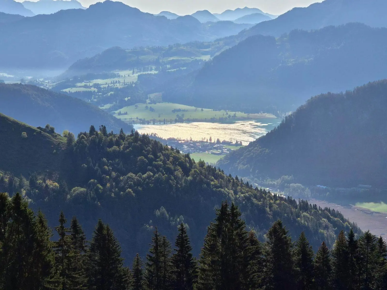 Ausblick vom Berg auf bewaldete Hügel, Täler und einen sonnenglitzernden See; dahinter staffeln sich Bergketten. | © DAV Markt Schwaben/Hubert Inhofer