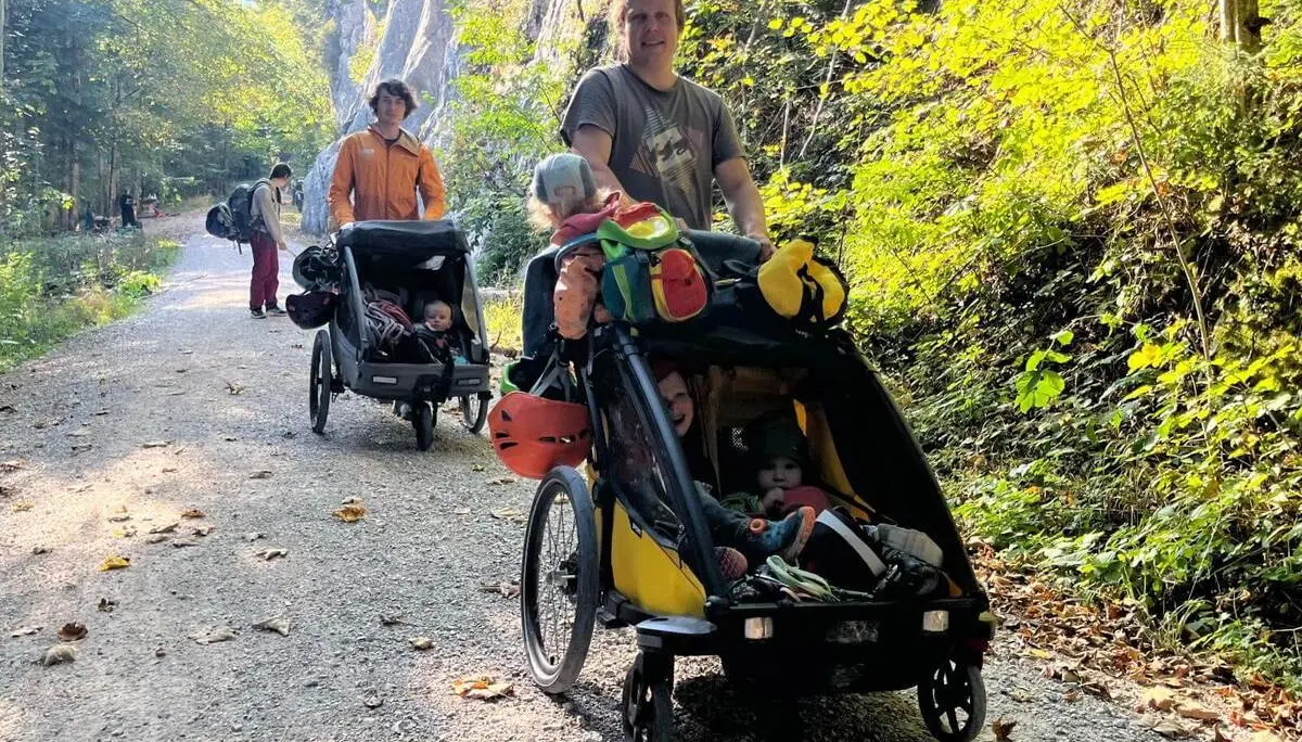 Familien mit Kindern sind auf einem breiten Waldweg zum Klettergarten unterwegs. Mehrere Kinder fahren in Anhängern mit. | © DAV Markt Schwaben/Lara Wolff