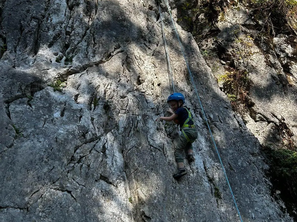 Ein Kind mit Kletterhelm und Gurt steigt an einer gesicherten Felswand empor. Das Seil verläuft oberhalb entlang der Route. | © DAV Markt Schwaben/Lara Wolff