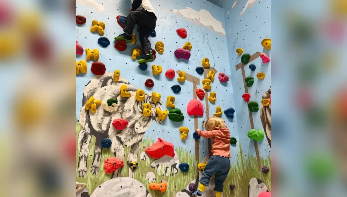 Zwei Kinder klettern an bunten Griffen vor einer Wand mit Tiermotiven; Schaf und Kuh im Hintergrund, Fokus auf Bewegung. | © DAV Markt Schwaben | Foto: Lara Wolff