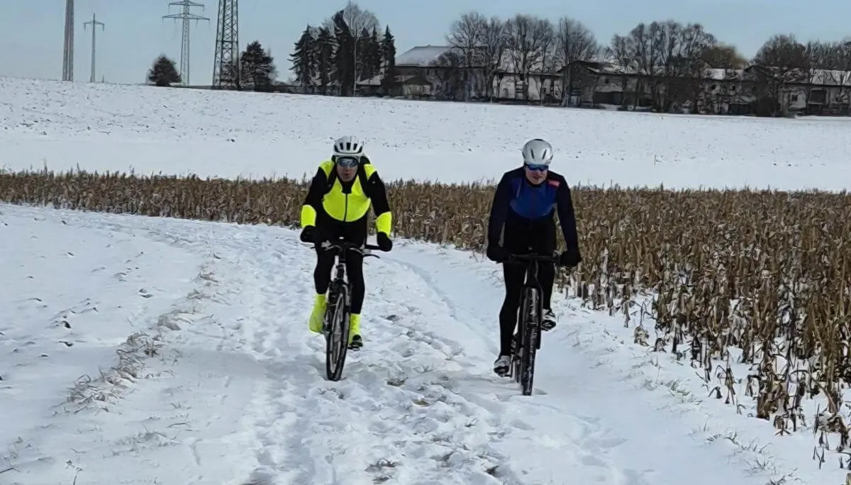 Zwei Radfahrer fahren nebeneinander auf verschneitem Feldweg; helle Kleidung hebt sich vom winterlichen Hintergrund ab. | © DAV Markt Schwaben | Foto: Miron Thoms