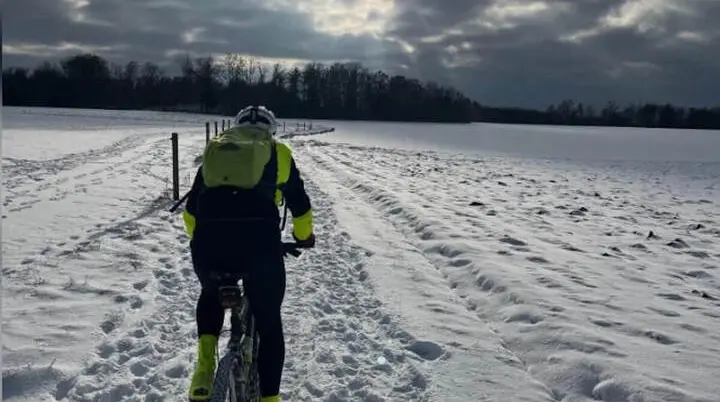 Eine Person fährt mit Mountainbike und gelber Jacke auf verschneitem Feldweg; Bäume und Sonne im wolkigen Hintergrund. | © DAV Markt Schwaben | Foto: Miron Thoms