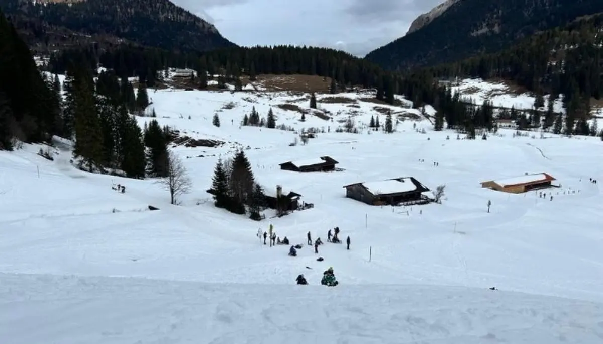 Verschneites Tal mit verstreuten Holzhütten; unten bewegen sich mehrere Menschen beim Aufstieg, umgeben von Wald und Bergkulisse unter grauem Himmel. | © DAV Markt Schwaben, Familiengruppe