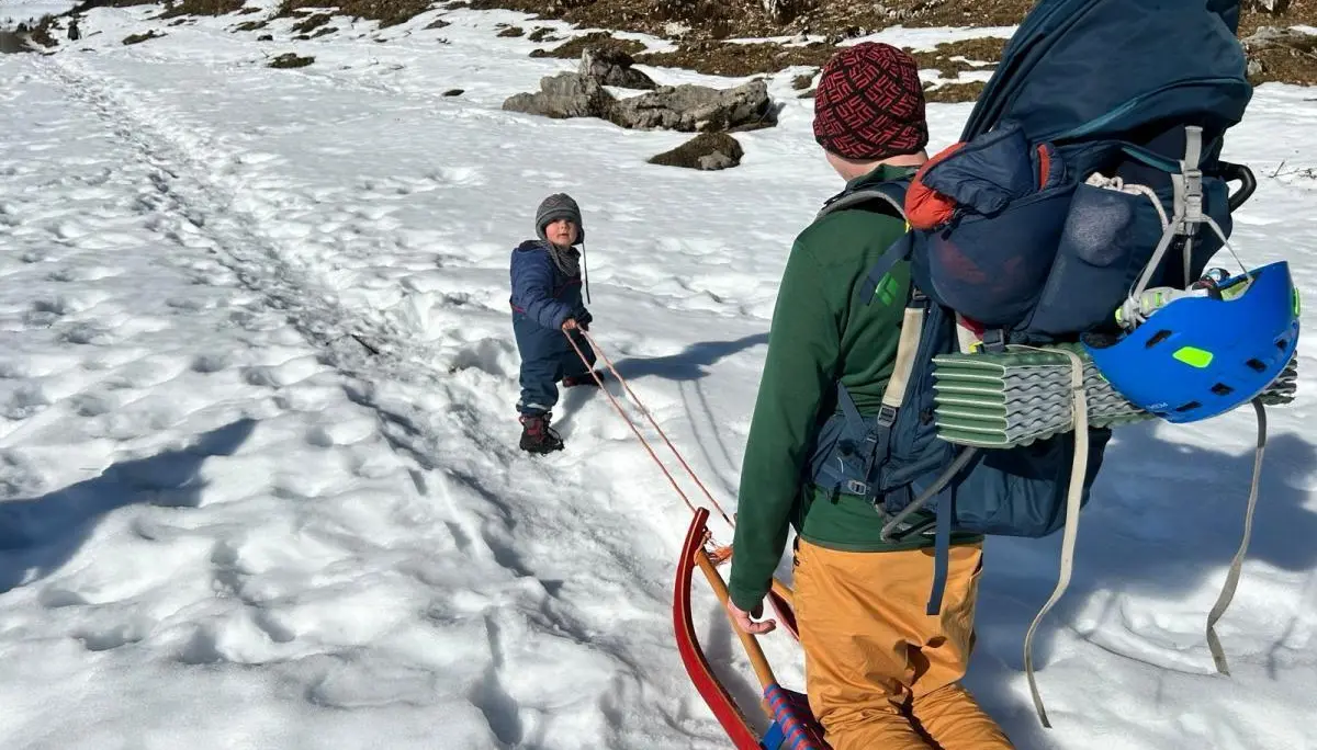 Ein Erwachsener zieht einen roten Schlitten über einen verschneiten Weg, ein Kind hält das Zugseil; beide tragen Winterkleidung, umgeben von Wald und Hügeln. | © DAV Markt Schwaben, Familiengruppe