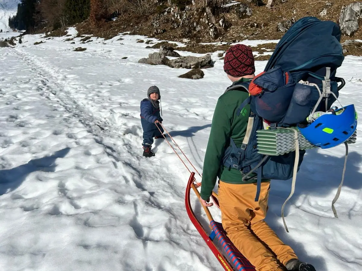Ein Erwachsener zieht einen roten Schlitten über einen verschneiten Weg, ein Kind hält das Zugseil; beide tragen Winterkleidung, umgeben von Wald und Hügeln. | © DAV Markt Schwaben, Familiengruppe