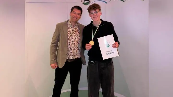 Zwei Personen stehen vor einem Banner mit der Aufschrift ‚Sportlerehrung Landkreis Ebersberg‘. Die geehrte Person trägt eine Goldmedaille um den Hals und hält eine Urkunde in den Händen.  | © Sportlerehrung Landkreis Ebersberg