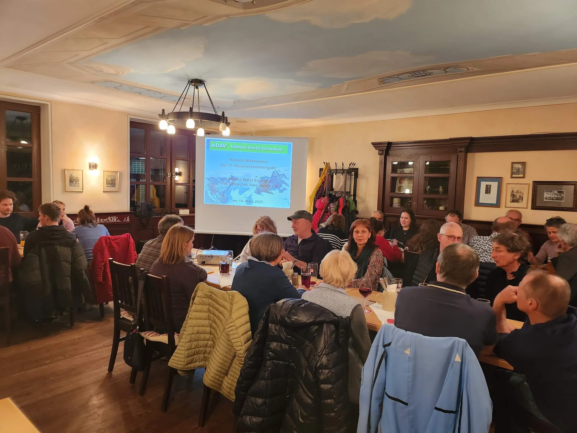 Mitglieder der DAV Sektion Markt Schwaben sitzen in einem Raum und schauen eine Präsentation auf einer Leinwand mit 'Herzlich Willkommen zur 17. Hauptversammlung DAV Sektion Markt Schwaben'. | © DAV Markt Schwaben | Foto Anna Thoms