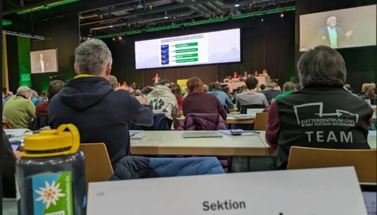Delegierte der DAV-Sektion Markt Schwaben sitzen im Konferenzsaal der Hauptversammlung in Passau. Im Vordergrund: Namensschild der Sektion und DAV-Wasserflasche. Auf der Bühne läuft eine Präsentation, Teilnehmende verfolgen konzentriert das Geschehen. | © DAV Markt Schwaben · Fotograf unbekannt
