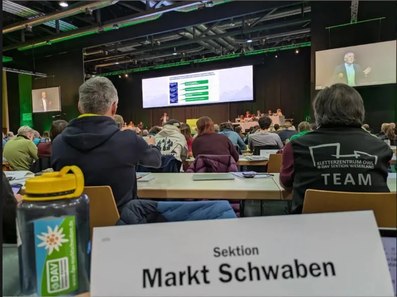 Delegierte der DAV-Sektion Markt Schwaben sitzen im Konferenzsaal der Hauptversammlung in Passau. Im Vordergrund: Namensschild der Sektion und DAV-Wasserflasche. Auf der Bühne läuft eine Präsentation, Teilnehmende verfolgen konzentriert das Geschehen. | © DAV Markt Schwaben · Fotograf unbekannt