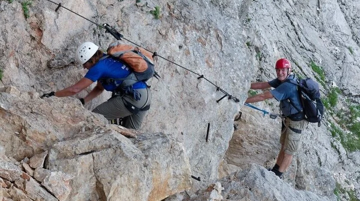 Zwei Kletternde mit Helm und Gurt steigen am Drahtseil entlang steiler Felswand; felsige Umgebung im Hintergrund. | © DAV Markt Schwaben | Foto Lutz Gründel