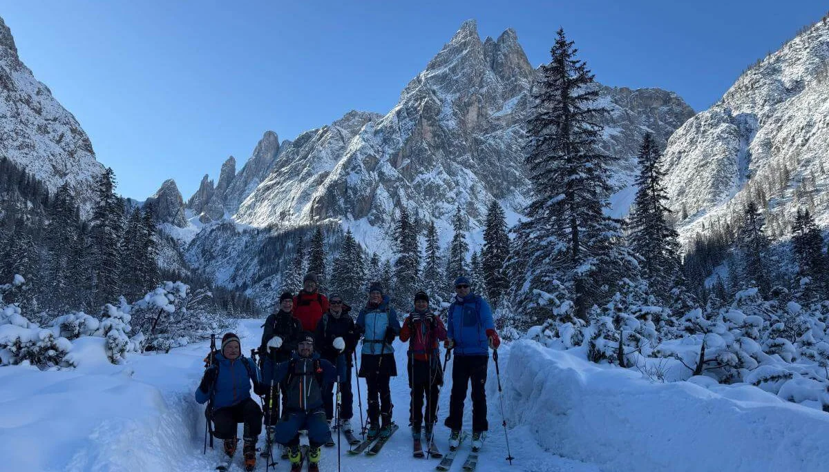 Acht Skitourengeher:innen stehen auf verschneitem Waldpfad im Fischleinbachtal vor sonnenbeschienenen Dolomitengipfeln. | © DAV Markt Schwaben · Foto: Benedikt Scheuerecke