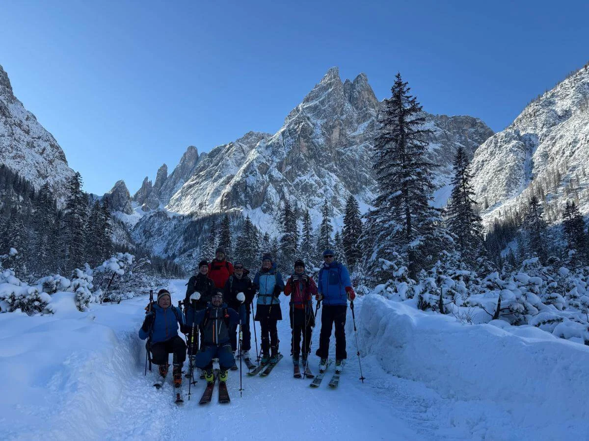 Acht Skitourengeher:innen stehen auf verschneitem Waldpfad im Fischleinbachtal vor sonnenbeschienenen Dolomitengipfeln. | © DAV Markt Schwaben · Foto: Benedikt Scheuerecke