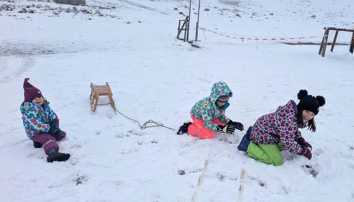 Drei Kinder in bunter Winterkleidung spielen im Schnee mit einem Holzschlitten, umgeben von einer Schneelandschaft.“ | © DAV Markt Schwaben