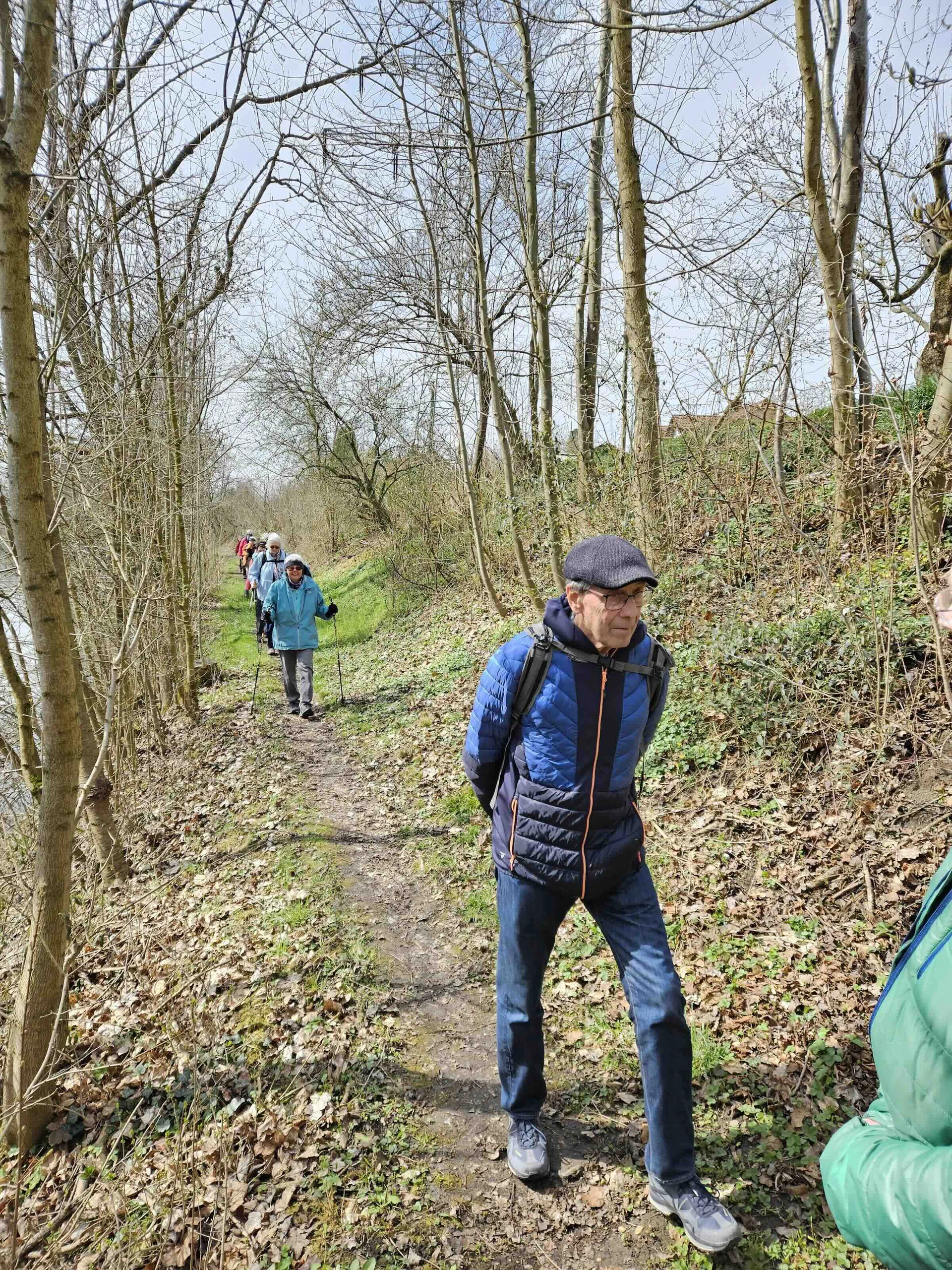 Eine Gruppe von Wanderern geht auf einem schmalen, unbefestigten Pfad durch ein bewaldetes Gebiet. Die Bäume haben überwiegend kahle Äste, was auf frühen Frühling oder späten Herbst hindeutet. Der Boden ist mit gefallenen Blättern und grünem Gras bedeckt. Die Wanderer tragen Outdoor-Kleidung, darunter Jacken und Rucksäcke. | © DAV Markt Schwaben