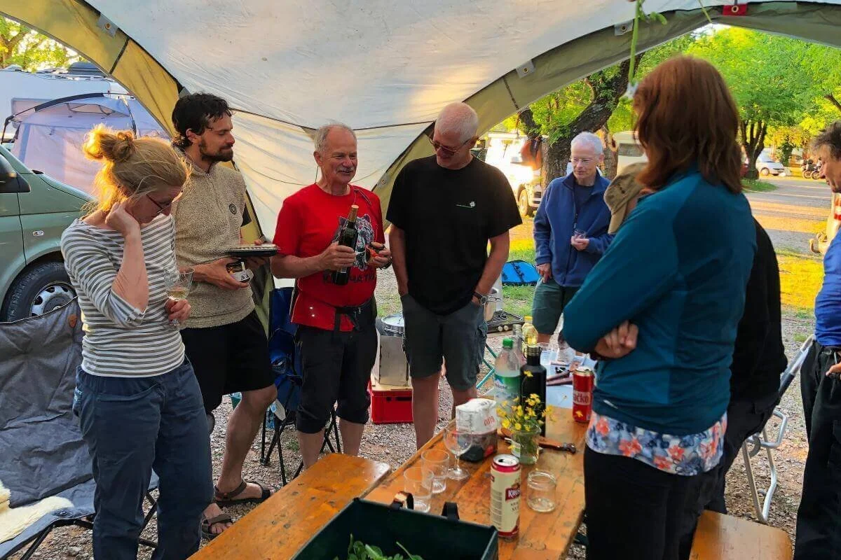 Acht Personen stehen oder sitzen am Tisch mit Getränken und Pflanze; Zelte und Van im Hintergrund. | © DAV Markt Schwaben | Foto Hildegard Petschik
