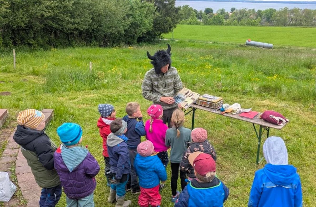 Eine Person mit einem Hut mit Hörnern zeigt einer Gruppe von Kindern draußen ein Buch. Sie stehen um einen Tisch mit Materialien. | © DAV Markt Schwaben | Foto: Familiengruppe