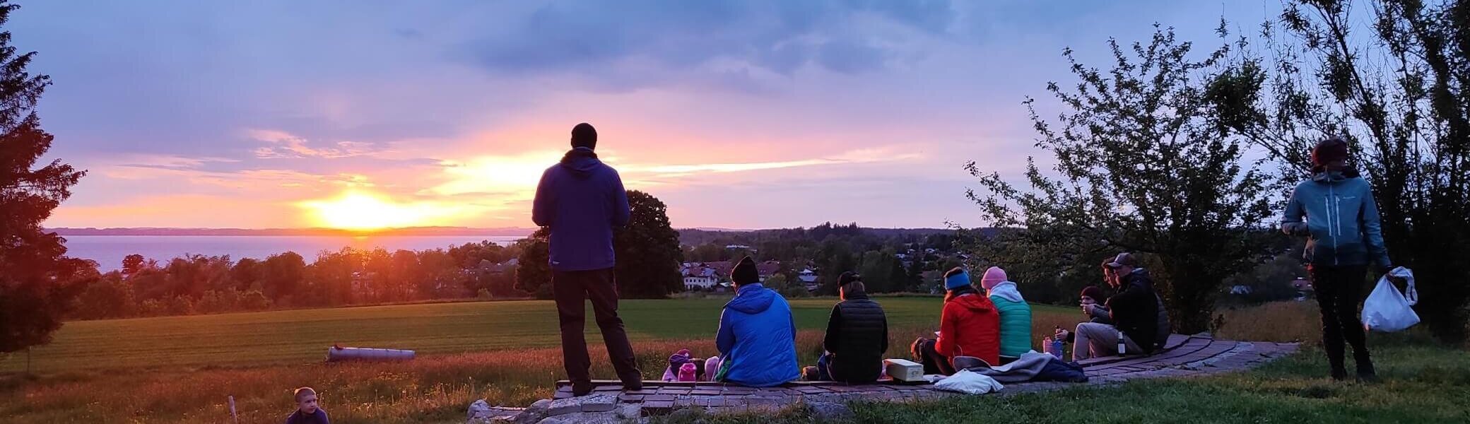 Eine Gruppe von Menschen sitzt auf einer Wiese und betrachtet den Sonnenuntergang. Der Himmel ist teilweise von Wolken bedeckt. | © DAV Markt Schwaben | Foto: Familiengruppe