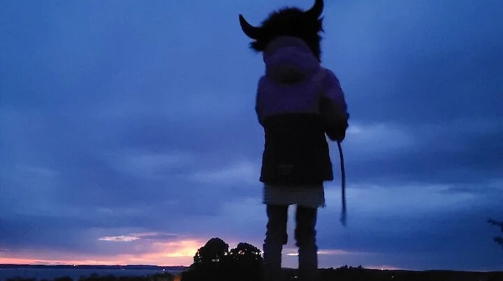 Eine Person mit einem Hut mit Hörnern steht bei Sonnenuntergang auf einem Hügel. Der Himmel leuchtet in warmen Blau- und Orangetönen. | © DAV Markt Schwaben | Foto: Familiengruppe