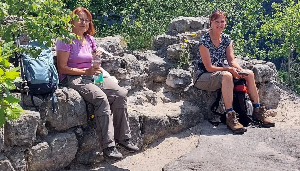 Zwei Personen sitzen auf Felsen mit gelben Blumen, umgeben von Grün, beide tragen Wanderausrüstung. | © DAV Markt Schwaben | Foto Lutz Gründel