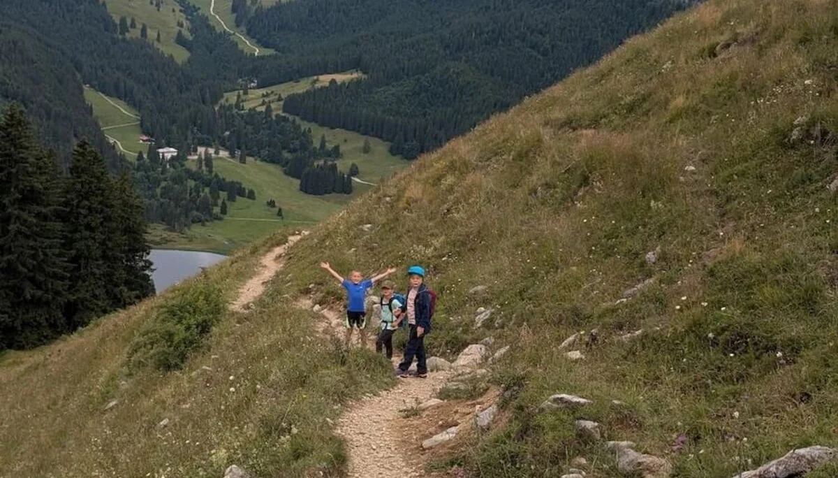 Drei Kinder wandern auf einem Bergpfad bei bewölktem Himmel. Sie gehen durch grünes Gelände mit Sicht auf umliegende Berge. | © DAV Markt Schwaben | Foto Familiengruppe