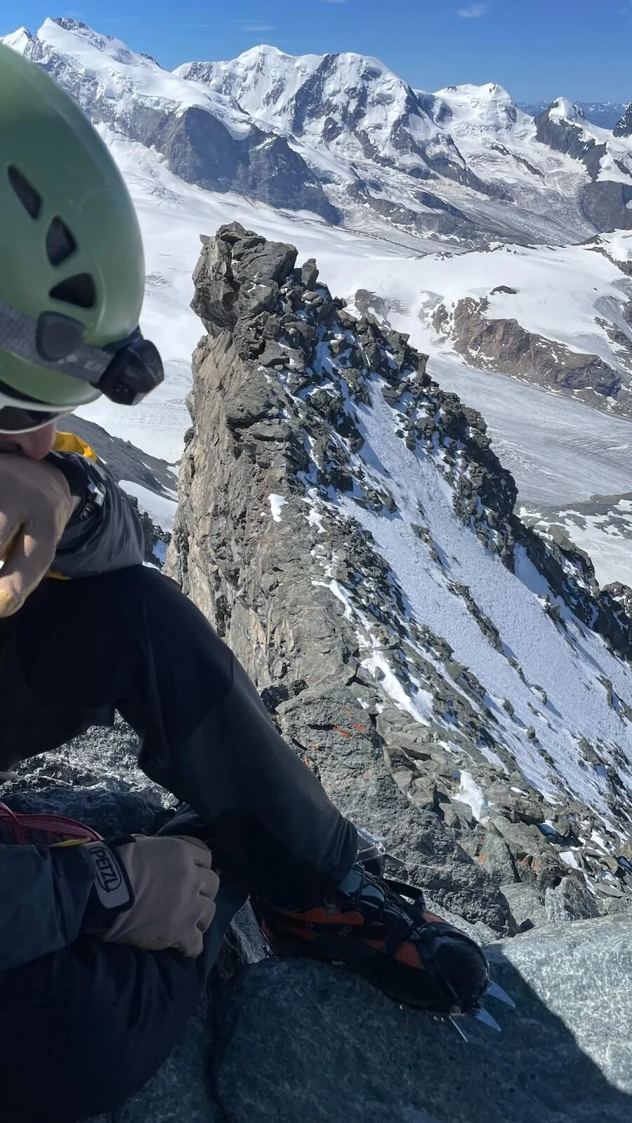 Ein Bergsteiger sitzt mit Helm und Steigeisen auf einem felsigen Grat der Westalpen – im Hintergrund Gletscher und klare Bergsicht. | © DAV Markt Schwaben | Christian Reischl