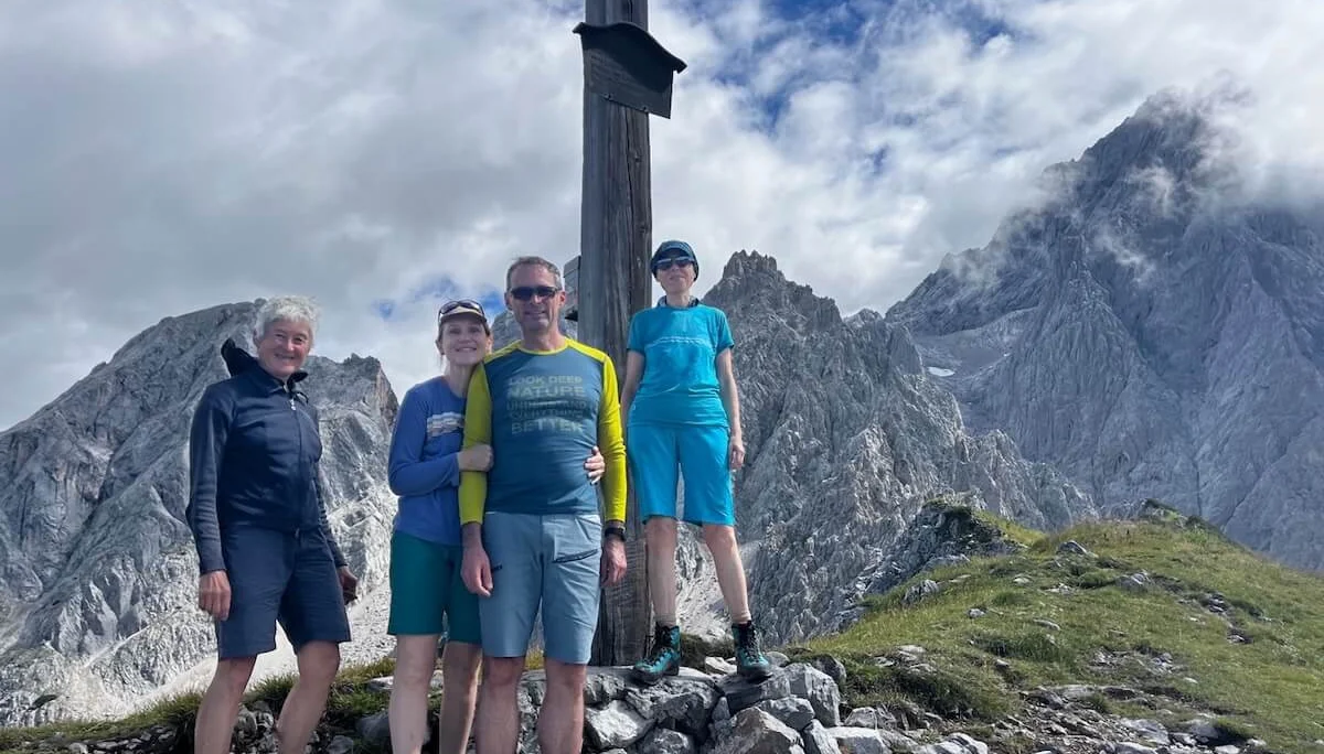 Vier Wandernde posieren vor einem Gipfelkreuz mit Blick auf felsige Bergspitzen unter teils bewölktem Himmel. | © DAV Markt Schwaben | Foto: Markus Sellmeier