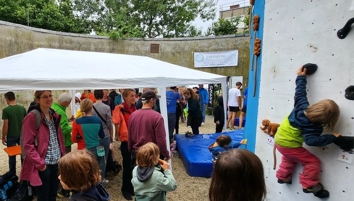 Zwei Kinder klettern gesichert an der Outdoor-Wand, umgeben von Besucher*innen unter einem weißen Pavillon. | © DAV Markt Schwaben | Foto Anna Thoms