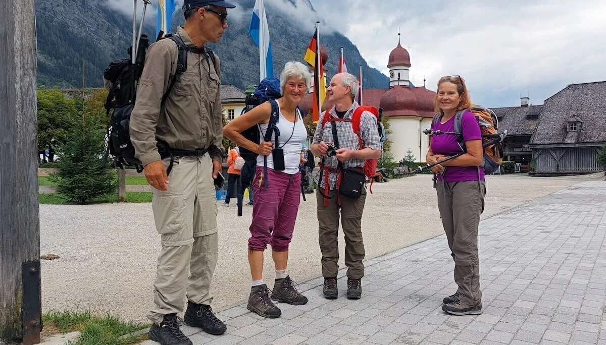 Vier Wanderer stehen mit Ausrüstung vor Kirche und Flaggen in St. Bartholomä, hinten bewaldeter Berg im Nebel. | © DAV Markt Schwaben · Foto: Lutz Gründel