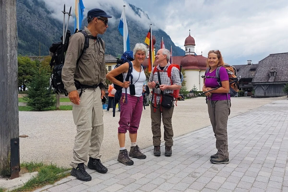 Vier Wanderer stehen mit Ausrüstung vor Kirche und Flaggen in St. Bartholomä, hinten bewaldeter Berg im Nebel. | © DAV Markt Schwaben · Foto: Lutz Gründel