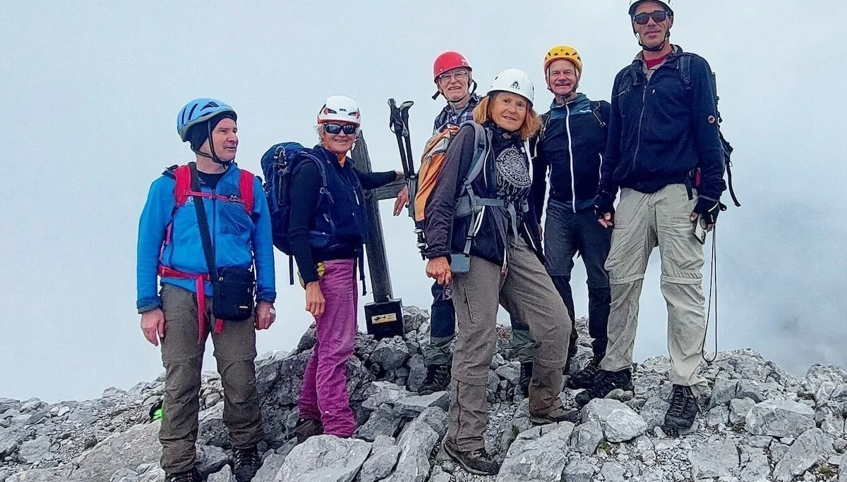 Sechs Bergsteiger stehen am Gipfelkreuz auf felsigem Grat, im Hintergrund Wolken und Bergstimmung. | © DAV Markt Schwaben · Foto: Lutz Gründel